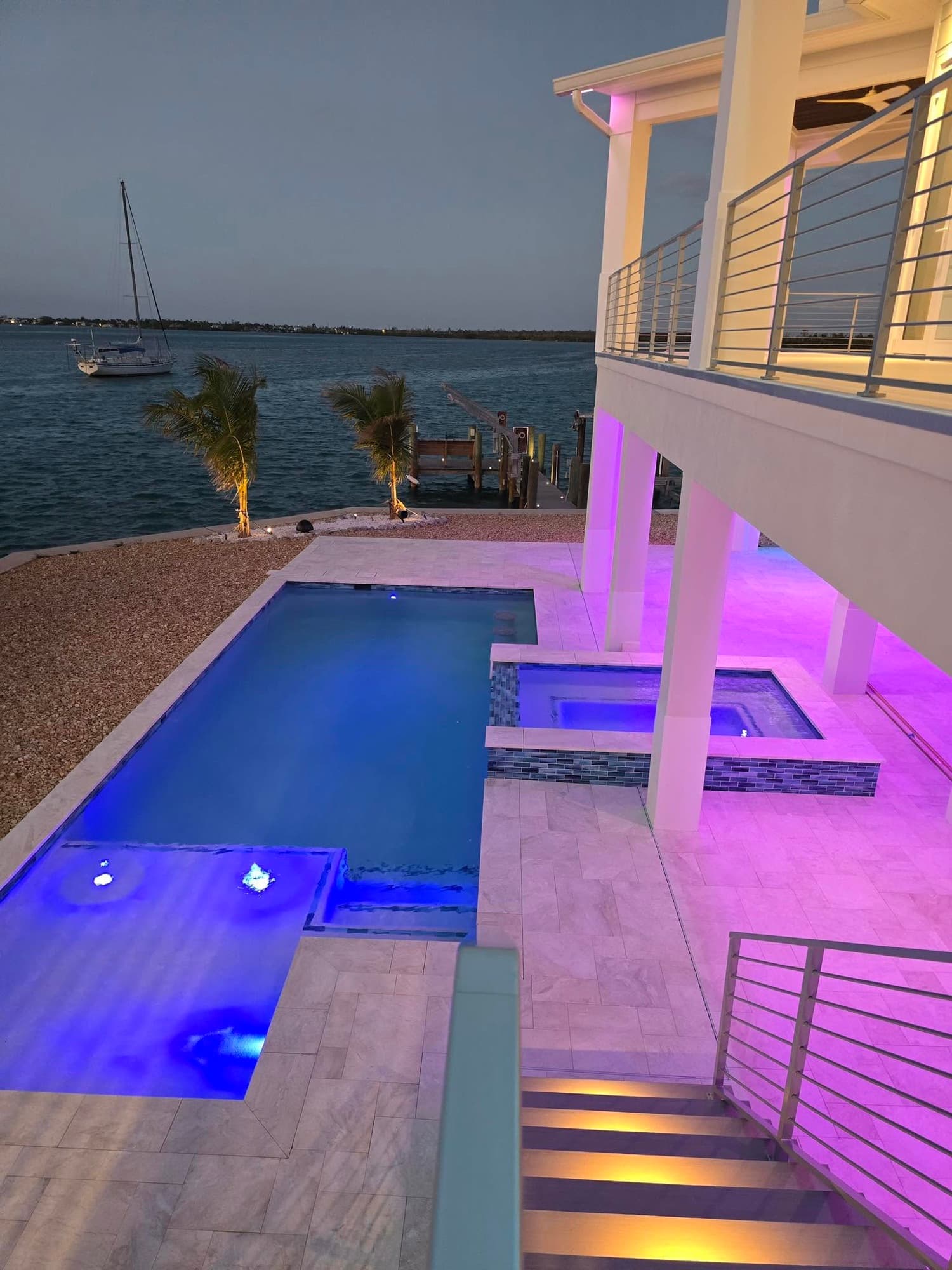 A pool deck at dusk with color-changing landscape lighting and a dock on the water behind it