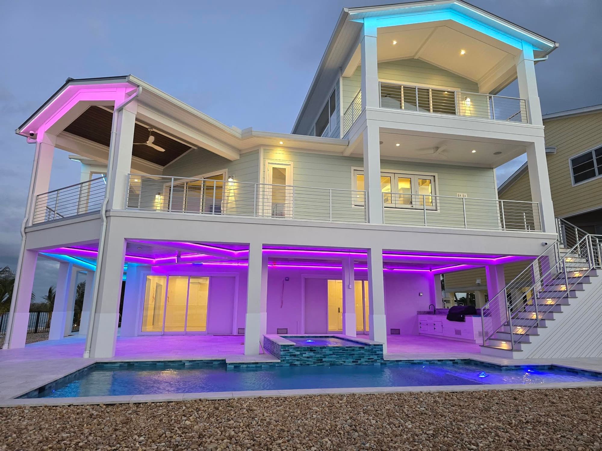 A three-story waterfront house at dusk with architectural LED lighting under the covered porches and a lit pool