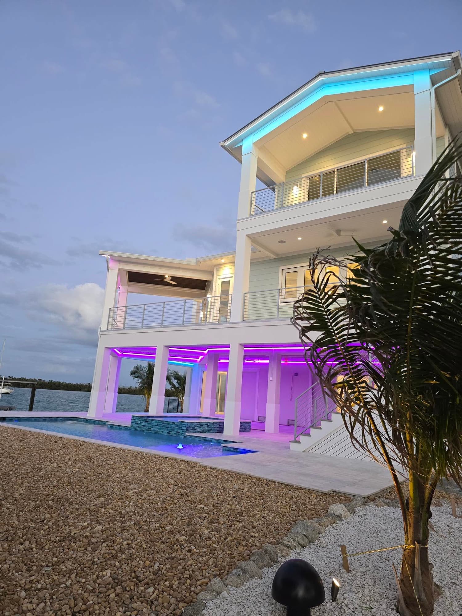 The same waterfront house from the side, palm tree in the foreground, exterior lighting on