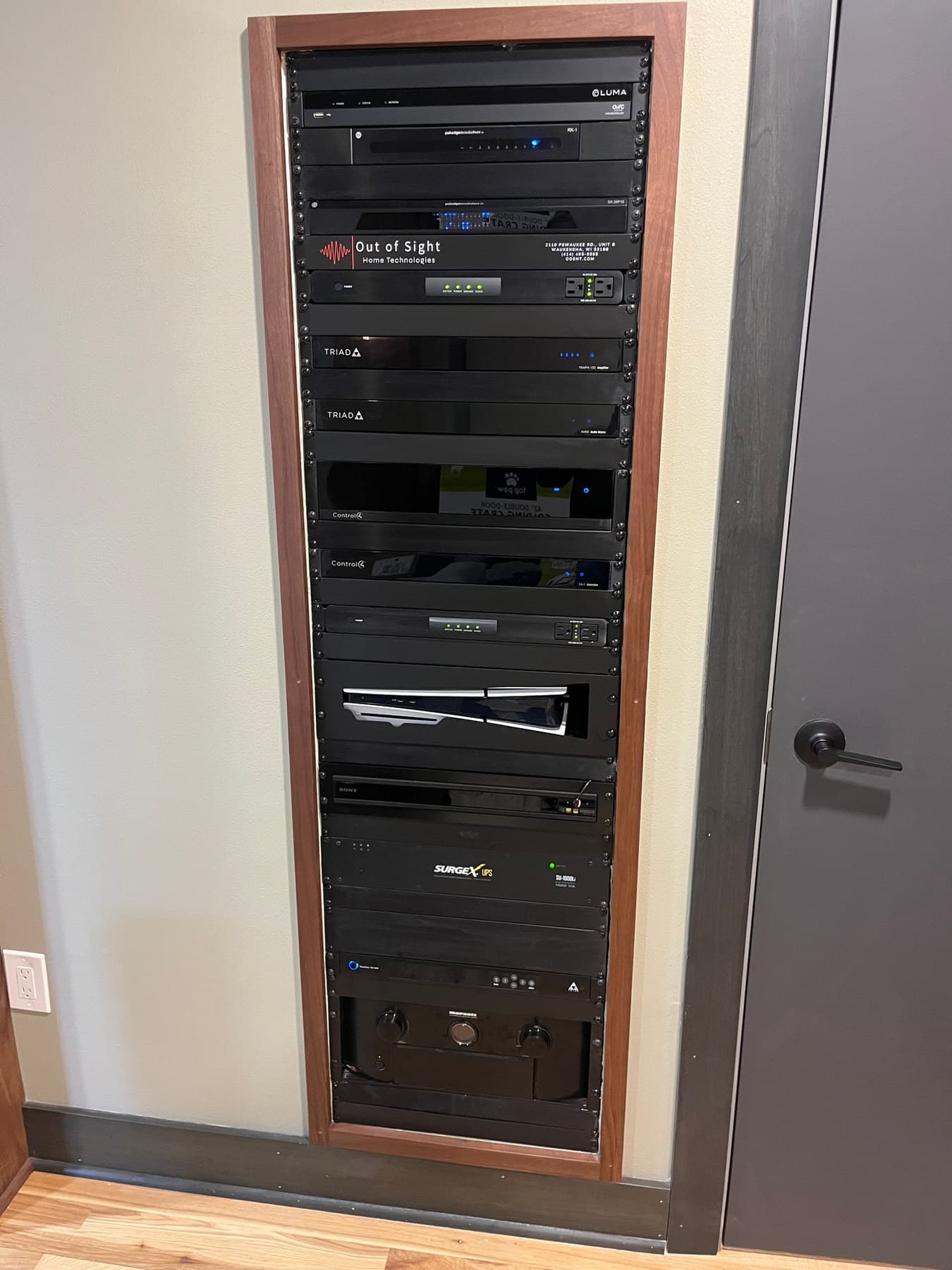 An installed equipment rack recessed into a walnut frame, carrying Control4, Triad, and Luma gear behind a labeled Out of Sight plate